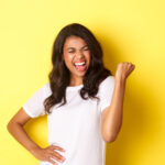 Portrait of happy african-american woman, triumphing and winning, making fist pump and shouting for joy, standing pleased against yellow background.